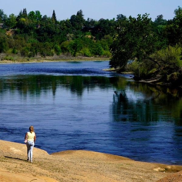 FILE – A woman walks along the banks of the American River flowing by the American River Parkway in Rancho Cordova, Calif., Friday, April 8, 2022. The U.S. Environmental Protection Agency has opened an investigation of how the California State Water Resources Control Board manages the state's water. Native American tribes and some environmental groups had filed a complaint alleging the state's policies discriminate against tribes and other communities of color. (AP Photo/Rich Pedroncelli, File)